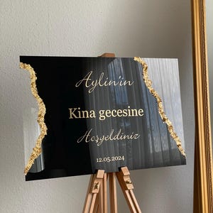 May include: Black and gold sign on a wooden easel. The sign has gold text that reads "Aylin'in Kina gecesine Hosgeldiniz 12.05.2024". The edges of the sign are decorated with gold leaf.