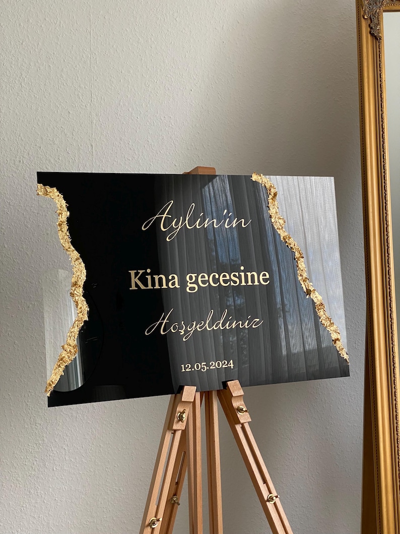 May include: Black and gold sign on a wooden easel. The sign has gold text that reads "Aylin'in Kina gecesine Hosgeldiniz 12.05.2024". The edges of the sign are decorated with gold leaf.
