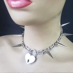 May include: Silver choker necklace with a heart-shaped padlock and sharp spikes. The necklace is made of metal and has a silver finish.