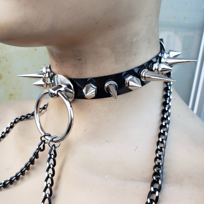 K&ouml;nnte beinhalten: Ein schwarzer Leder-Choker mit silbernen Spikes und einem silbernen Ring, der an einer silbernen Kette befestigt ist.