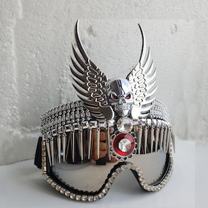 Peut inclure: Lunettes de ski argentées et noires avec un motif de crâne et d'ailes. Les lunettes ont un bijou rouge sur le crâne et un bijou rouge sur le devant des lunettes. Les lunettes sont décorées de strass et de pointes.