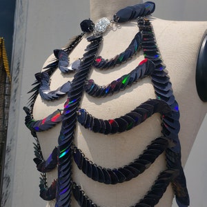 K&ouml;nnte beinhalten: Eine schwarze, schimmernde Kettenkette im Kettenhemd-Stil mit einem silbernen Verschluss. Die Kette besteht aus kleinen, &uuml;berlappenden Schuppen, die einen schimmernden Effekt erzeugen.