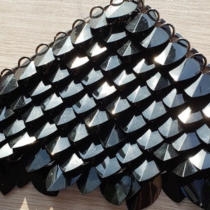 May include: Close-up of a section of black scale armour. The scales are shaped like elongated teardrops and are connected by small, dark rings. The surface of the scales reflects light, creating highlights and shadows. The armour is likely for cosplay or historical reenactment.