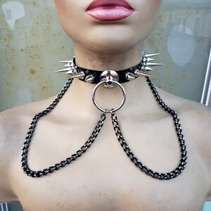 K&ouml;nnte beinhalten: Ein schwarzes Lederhalsband mit silbernen Spikes und einem silbernen Ring. Das Halsband ist an einer silbernen Kette mit schwarzer Oberfl&auml;che befestigt.