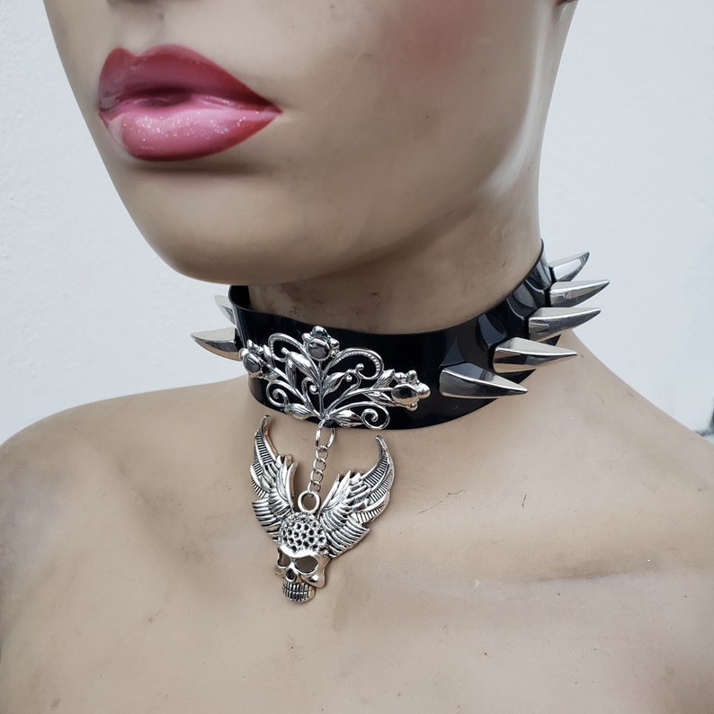 May include: Black leather choker with silver spikes and a dangling skull pendant with wings. The pendant features a floral design.