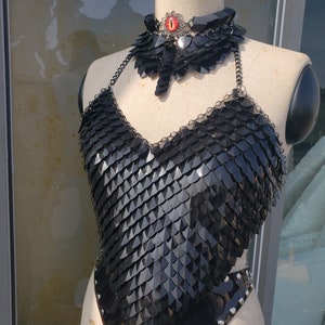 Op de afbeelding: Een zwart kettinghemd top met een zilveren oog hanger ketting. De top is gemaakt van kleine, overlappende metalen schubben en heeft een halter halslijn.