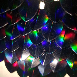 Peut inclure: Gros plan sur une section de matériau irisé en forme d'écailles. Les écailles sont d'une couleur sombre et réfléchissante, avec des teintes arc-en-ciel vibrantes de rouge, de vert et de bleu. Les écailles se chevauchent, créant un effet texturé et superposé.