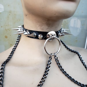 K&ouml;nnte beinhalten: Ein schwarzes Lederhalsband mit silbernen Spikes und einem silbernen Ring. Das Halsband ist an einer silbernen Kette befestigt.