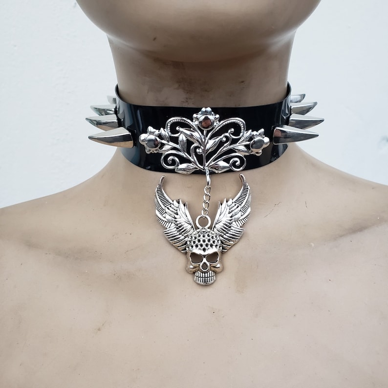 May include: Black leather choker with silver spikes and a silver skull pendant with wings. The pendant is attached to a silver chain with a floral design.