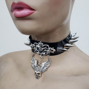 May include: A black leather choker with silver spikes and a silver pendant featuring a skull with wings and a floral design.