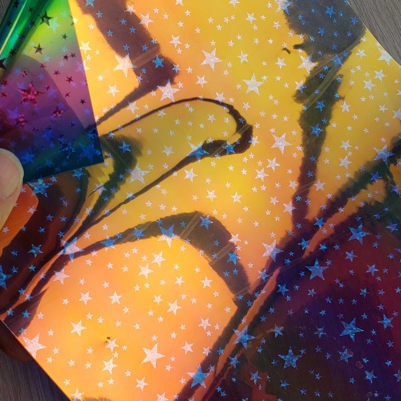 May include: A sheet of iridescent holographic paper with a pattern of white stars on a yellow, orange, and purple background.