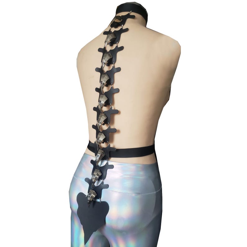 K&ouml;nnte beinhalten: Ein schwarzes, r&uuml;ckenwirbelf&ouml;rmiges K&ouml;rpergeschirr mit silberfarbenen Akzenten. Das Geschirr verf&uuml;gt &uuml;ber einen Choker, einen G&uuml;rtel und ein vertikales R&uuml;ckgratdesign mit beweglichen Metallteilen. Das Design wird auf einer Schaufensterpuppe mit silbernen Leggings getragen.