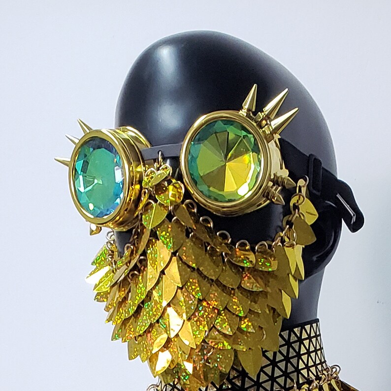 Pu&ograve; includere: Una maschera in stile steampunk dorata e verde con lenti a iridescenza e una frangia a maglia dorata. La maschera &egrave; indossata su una testa di manichino nera.