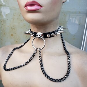 K&ouml;nnte beinhalten: Ein schwarzer Leder-Choker mit silbernen Spikes und einer silbernen Kette, die an einem silbernen Ring befestigt ist.