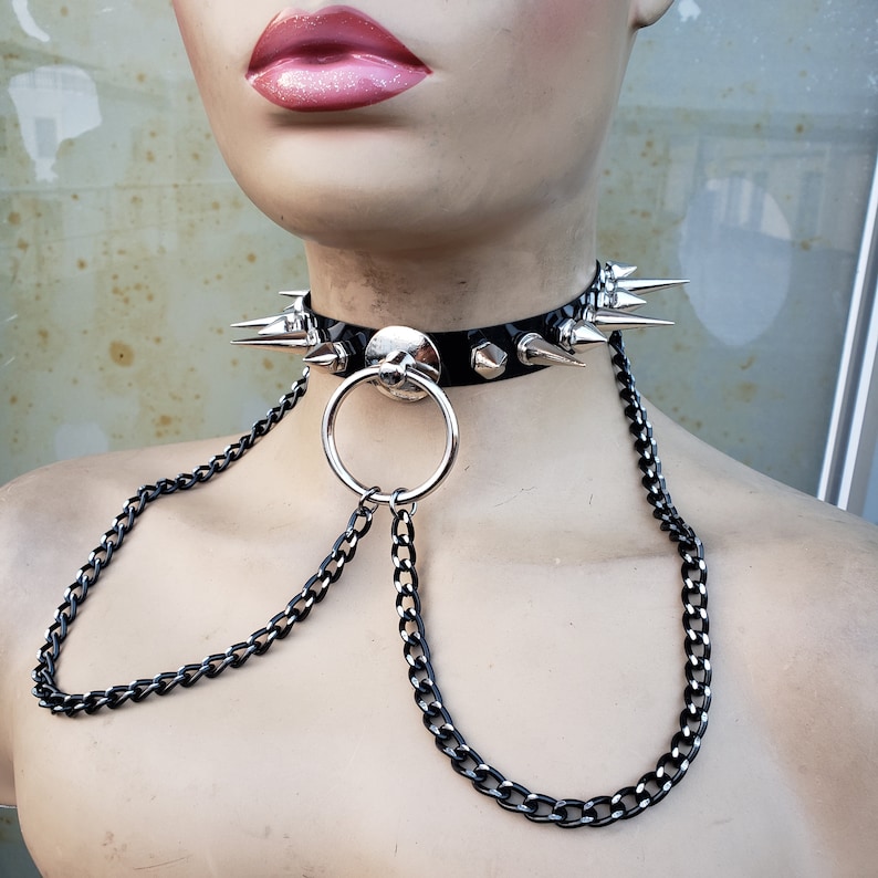 K&ouml;nnte beinhalten: Ein schwarzer Leder-Choker mit silbernen Spikes und einer silbernen Kette, die an einem silbernen Ring befestigt ist.