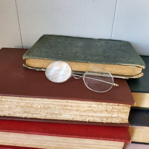 May include: A pair of antique silver-toned reading glasses with round lenses on top of a stack of books. The top book is green with gold lettering and the bottom book is red.