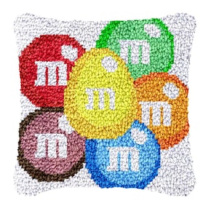 Puede incluir: Una almohada blanca con seis parches redondos de colores, cada uno con la letra "M" en blanco. Los parches son rojo, amarillo, verde, naranja, marrón y azul.