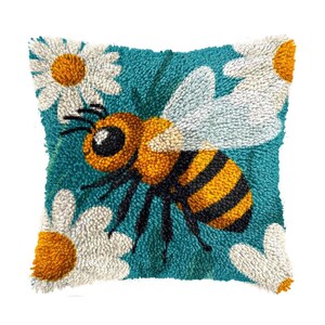 May include: Square decorative pillow with a teal background featuring a large bee with yellow and black stripes, white wings, and large black eyes. White daisies with yellow centers are scattered around the bee. The pillow is made of a textured material.