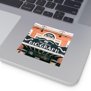 Könnte beinhalten: Ein Aufkleber mit dem Biograph Theater in Chicago. Der Aufkleber zeigt das Markenzeichen des Theaters mit dem Namen "BIOGRAPH" in großen weißen Buchstaben. Das Gebäude besteht aus roten Ziegeln und hat eine dekorative Fassade.