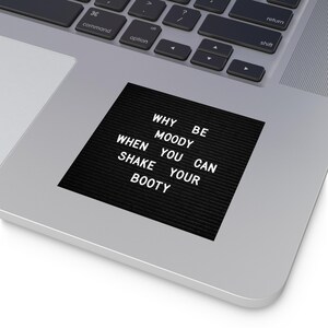 May include: A black sticker with white text that reads "WHY BE MOODY WHEN YOU CAN SHAKE YOUR BOOTY." The sticker is on a laptop, with the keyboard visible in the background.