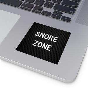 May include: A black square sticker with the words "SNORE ZONE" in white, placed on a silver laptop. The sticker has a textured surface, and the text is in a bold, sans-serif font. The laptop keyboard is visible in the background.