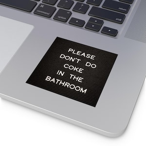 May include: A black sticker with white text that reads "PLEASE DON'T DO COKE IN THE BATHROOM" is placed on a silver laptop. The sticker is rectangular and has a textured surface. The laptop keyboard is visible in the background.