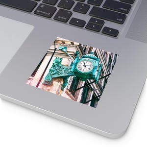 Könnte beinhalten: Ein Aufkleber mit einer Vintage-Uhr in Türkis. Die Uhr ist an einer Gebäudefassade angebracht und hat ein weißes Zifferblatt mit schwarzen Zeigern und römischen Ziffern. Die Uhr ist von aufwendigen, dekorativen Metallarbeiten umgeben.