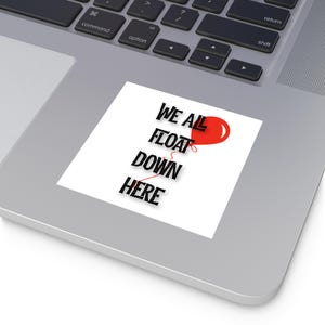 May include: A white sticker featuring the phrase "WE ALL FLOAT DOWN HERE" in black, with a red balloon graphic. The sticker is placed on a laptop.