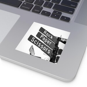 May include: Black and white sticker featuring street signs pointing in different directions. The signs read "Rock", "Paper", and "Scissors". The sticker is placed on a laptop.