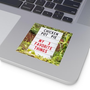 May include: A white sign with the text "CHICKEN POT PIE MY 3 FAVORITE THINGS" in black and red letters. The sign is surrounded by green foliage and sits on a wooden fence.