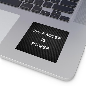Può includere: Un adesivo quadrato nero con le parole "CHARACTER IS POWER" in bianco, posto su un laptop argentato. L'adesivo ha una superficie strutturata e il testo è centrato.