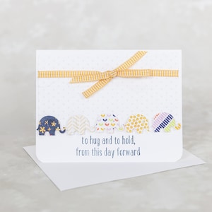 May include: A white card with a yellow ribbon tied in a bow. The card has a line of five colorful elephants with the text "to hug and to hold, from this day forward".