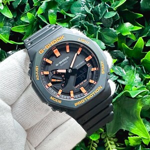 May include: A black G-Shock watch with orange accents. The watch face has a digital display and an analog display. The watch has a black band and a black case. The watch is on a white surface.