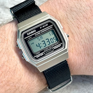 Pu&ograve; includere: Un orologio digitale Casio F-91W argentato con un cinturino in nylon nero. Il quadrante dell'orologio mostra l'ora, la data e le impostazioni della sveglia. L'orologio &egrave; resistente all'acqua.