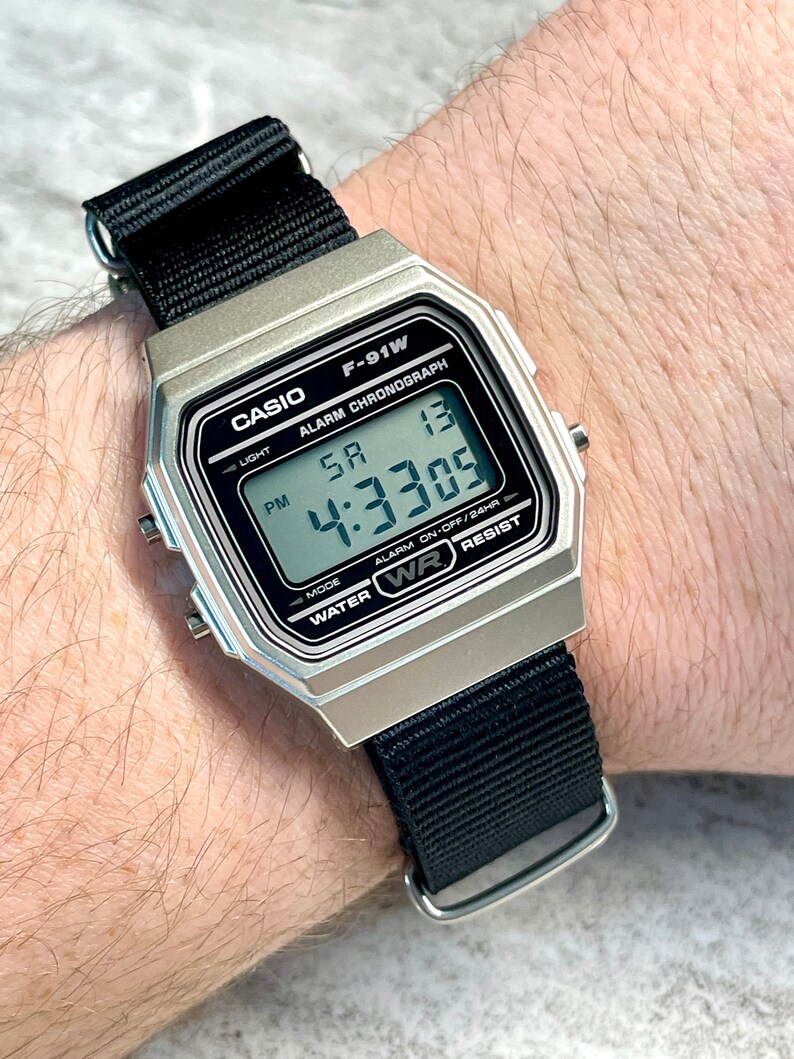 Pu&ograve; includere: Un orologio digitale Casio F-91W argentato con un cinturino in nylon nero. Il quadrante dell'orologio mostra l'ora, la data e le impostazioni della sveglia. L'orologio &egrave; resistente all'acqua.
