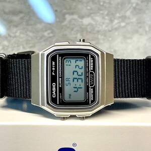 Pu&ograve; includere: Un orologio digitale Casio F-91W argentato con un cinturino in nylon nero. Il quadrante dell'orologio mostra l'ora, la data e le impostazioni della sveglia. L'orologio &egrave; resistente all'acqua.