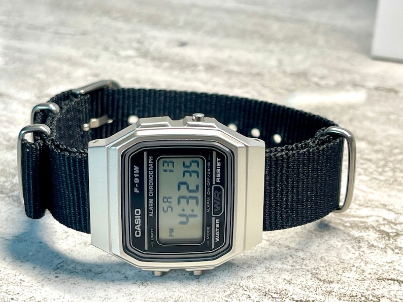 Pu&ograve; includere: Un orologio digitale Casio F-91W argentato con un cinturino in nylon nero. Il quadrante dell'orologio mostra l'ora, la data e le impostazioni della sveglia. L'orologio &egrave; resistente all'acqua.