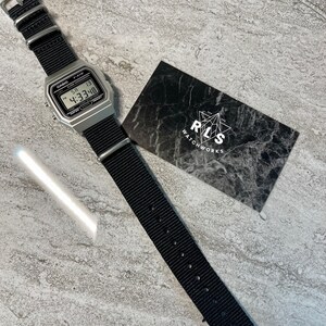 Pu&ograve; includere: Un orologio digitale Casio F-91W argentato con un cinturino in nylon nero. Il quadrante dell'orologio mostra l'ora come 4:33:48. L'orologio &egrave; su una superficie grigia con una carta nera che dice "RLS WATCHWORKS" in testo bianco.