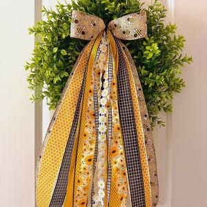 May include: A decorative wreath with a green base and a large bow featuring bee and honeycomb patterns. The wreath is adorned with multiple ribbons in yellow, black, and white, with sunflower and daisy designs. The wreath is hanging on a white door.