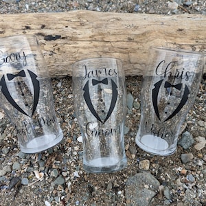 May include: Three clear glass pint glasses with black tuxedo designs and personalized names. The glasses say "Gary Best Man", "James Groom", and "Chris Usher".