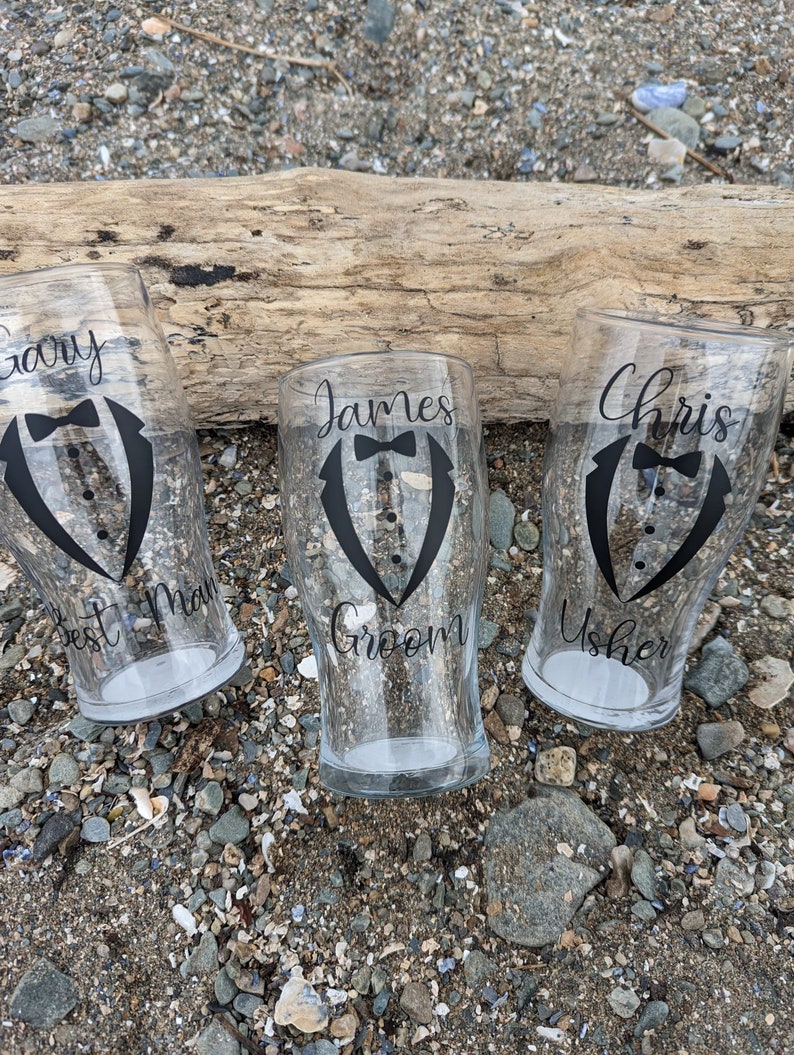 May include: Three clear glass pint glasses with black tuxedo designs and personalized names. The glasses say "Gary Best Man", "James Groom", and "Chris Usher".