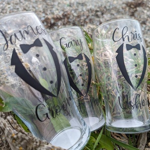 May include: Three clear glass cups with black vinyl decals. The decals feature a tuxedo with a bow tie and the names "James Groom", "Gary Best Man", and "Chris Usher".