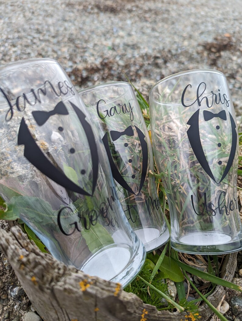 May include: Three clear glass cups with black vinyl decals. The decals feature a tuxedo with a bow tie and the names "James Groom", "Gary Best Man", and "Chris Usher".