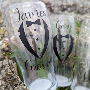 May include: Three clear glass pint glasses with black vinyl decals. The glasses are decorated with a tuxedo graphic and the names "James Groom", "Gary", and "Best Man".