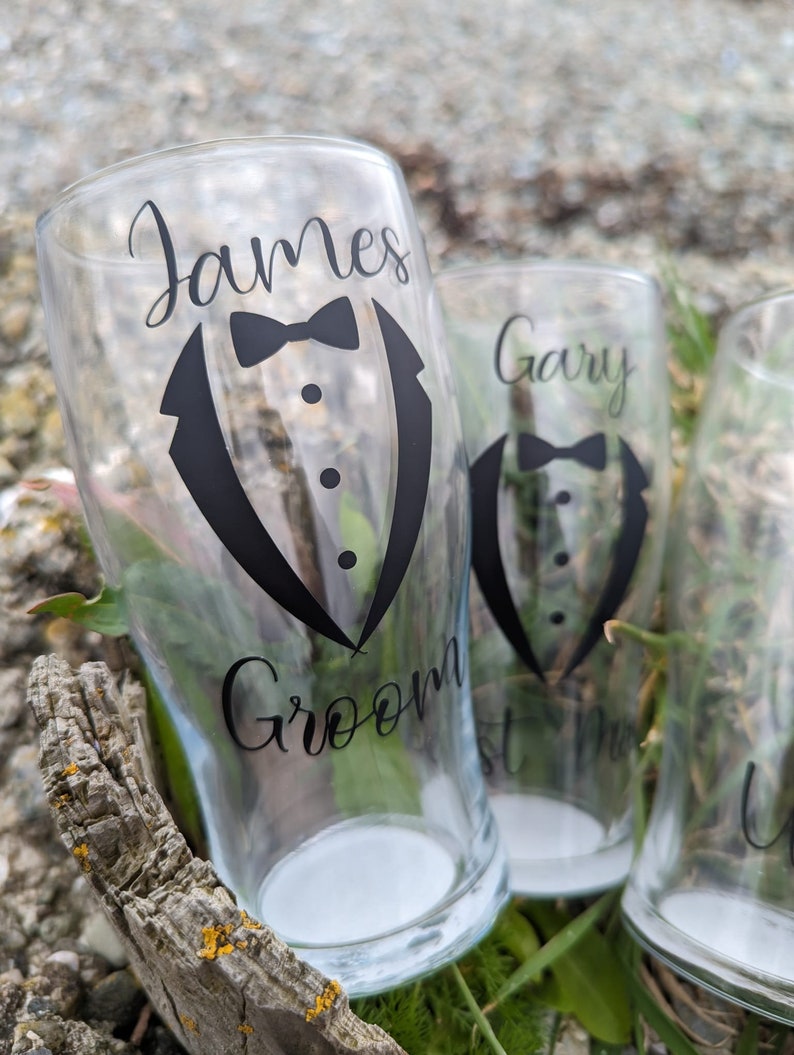 May include: Three clear glass pint glasses with black vinyl decals. The glasses are decorated with a tuxedo graphic and the names "James Groom", "Gary", and "Best Man".