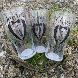 May include: Three clear glass pint glasses with black tuxedo designs and names printed on them. The glasses read "James Groom", "Gary Best Man", and "Chris Usher".