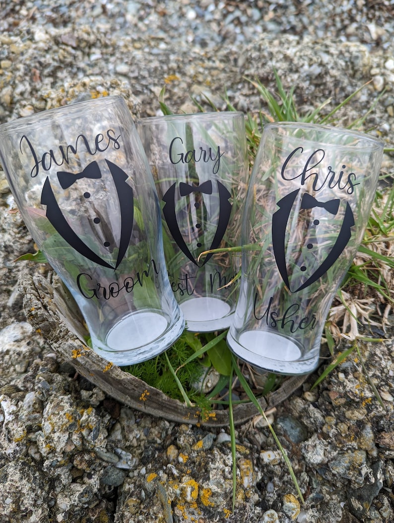 May include: Three clear glass pint glasses with black tuxedo designs and names printed on them. The glasses read "James Groom", "Gary Best Man", and "Chris Usher".