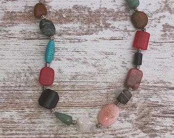 May include: A necklace made of various colored stones, including green, red, pink, white, and brown. The stones are strung on a silver chain with silver beads between each stone.