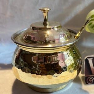 May include: A gold hammered metal sugar bowl with a lid and a small spoon. The bowl has a textured surface and a rounded shape. The spoon is made of metal and has a rounded handle.