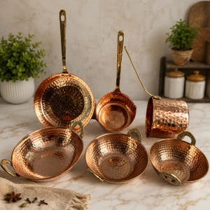 May include: A set of copper-toned kitchen utensils with a hammered finish. The collection features a large pan, a ladle, a small pot, a cylindrical container, and two bowls. The handles and rims have a gold finish. The items are displayed on a marble surface.